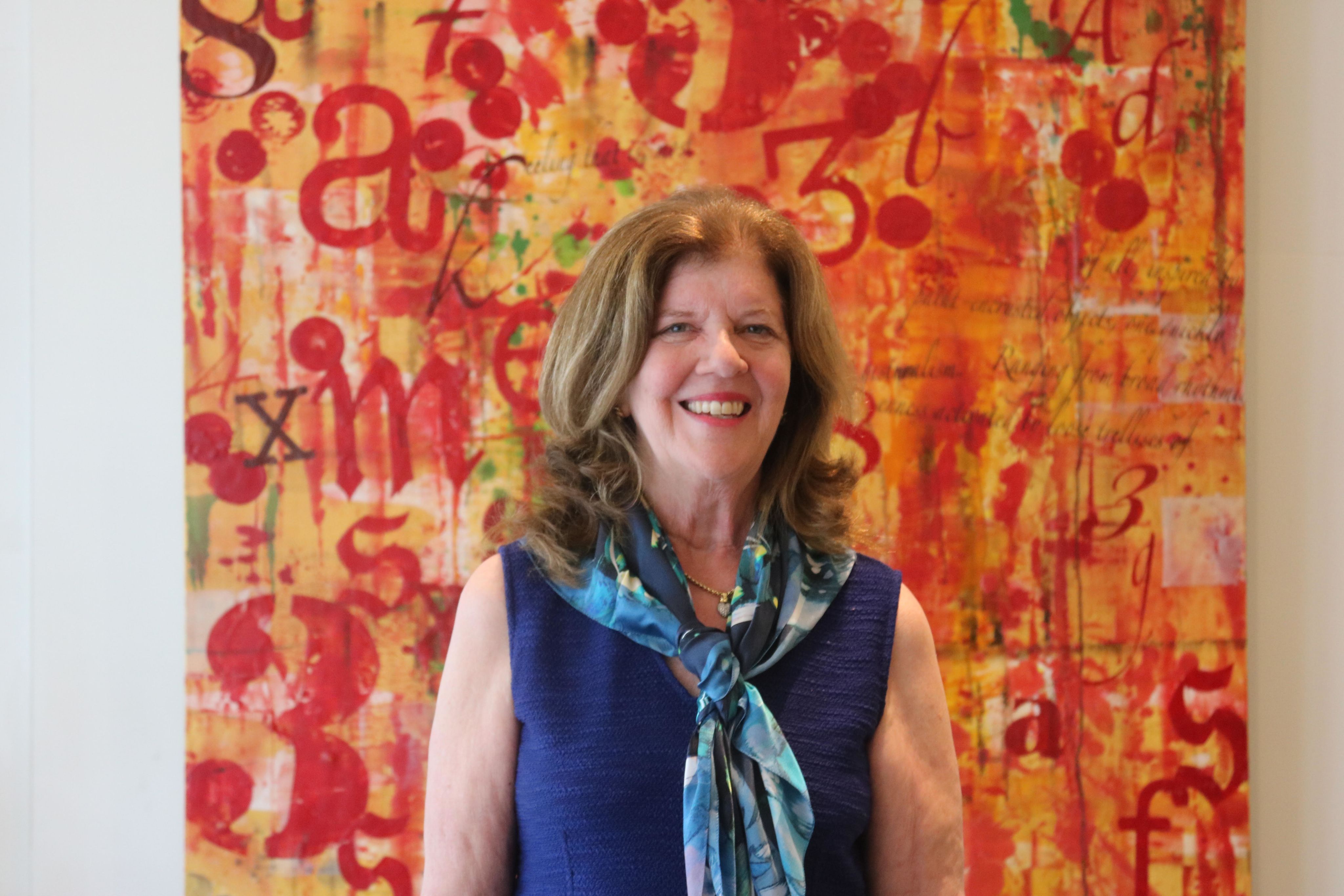 A woman standing in front of a painting with a warm smile on her face. She is wearing a necklace, adding a touch of elegance to her look.