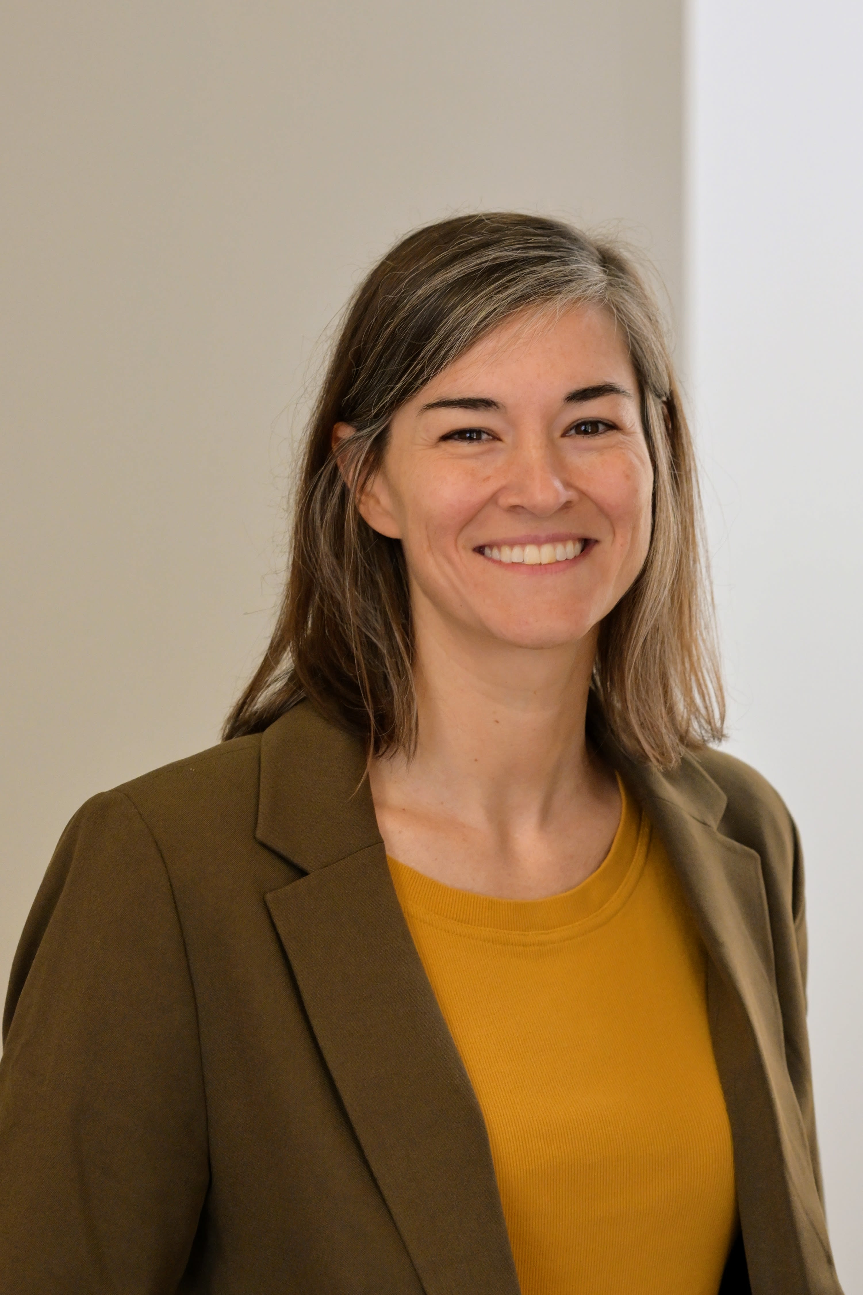 A woman in a yellow top and brown blazer standing in front of a white wall, smiling confidently.