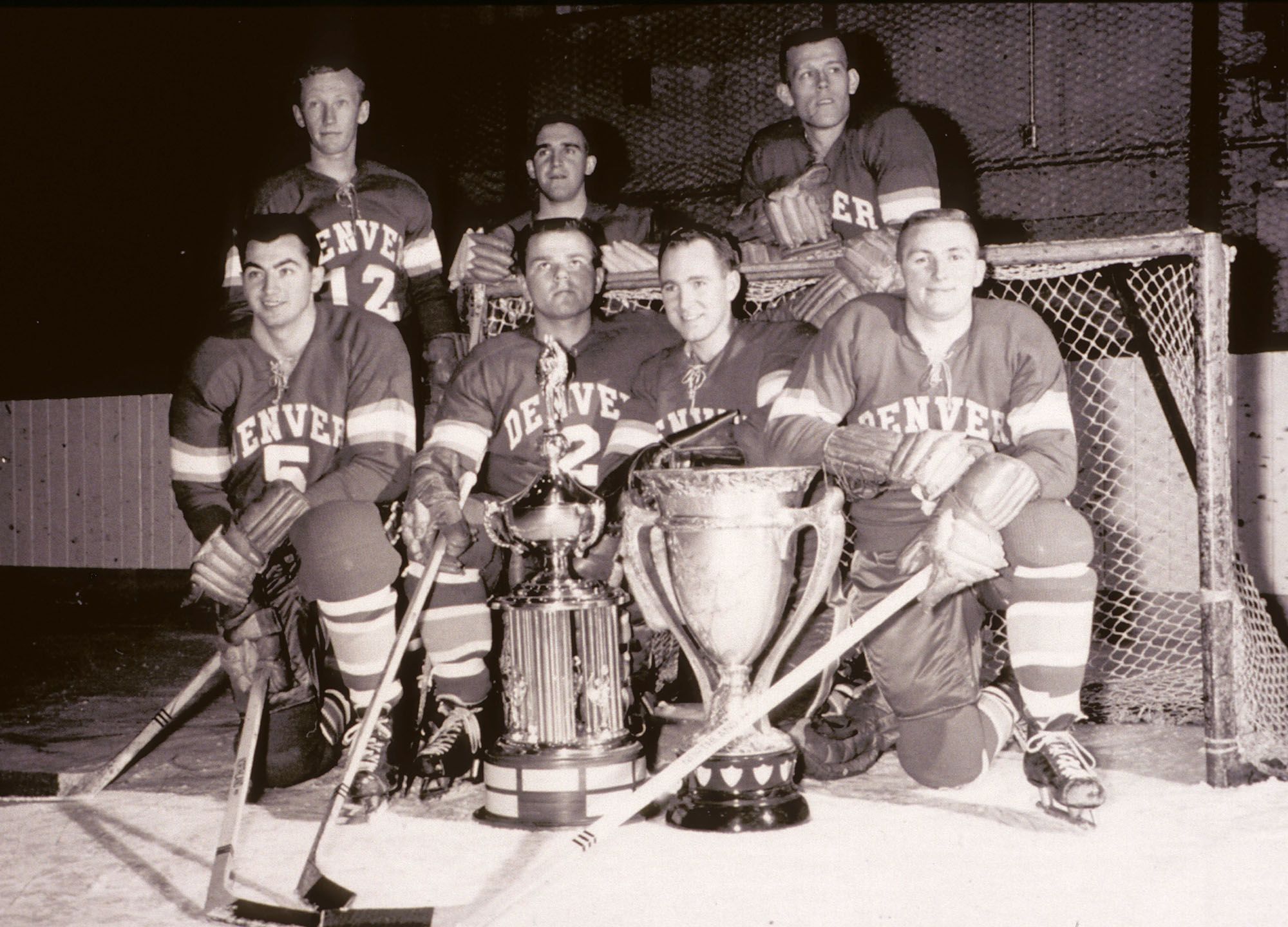 7 DU hockey players pose in front of a goalie net with hockey sticks and a trophy.