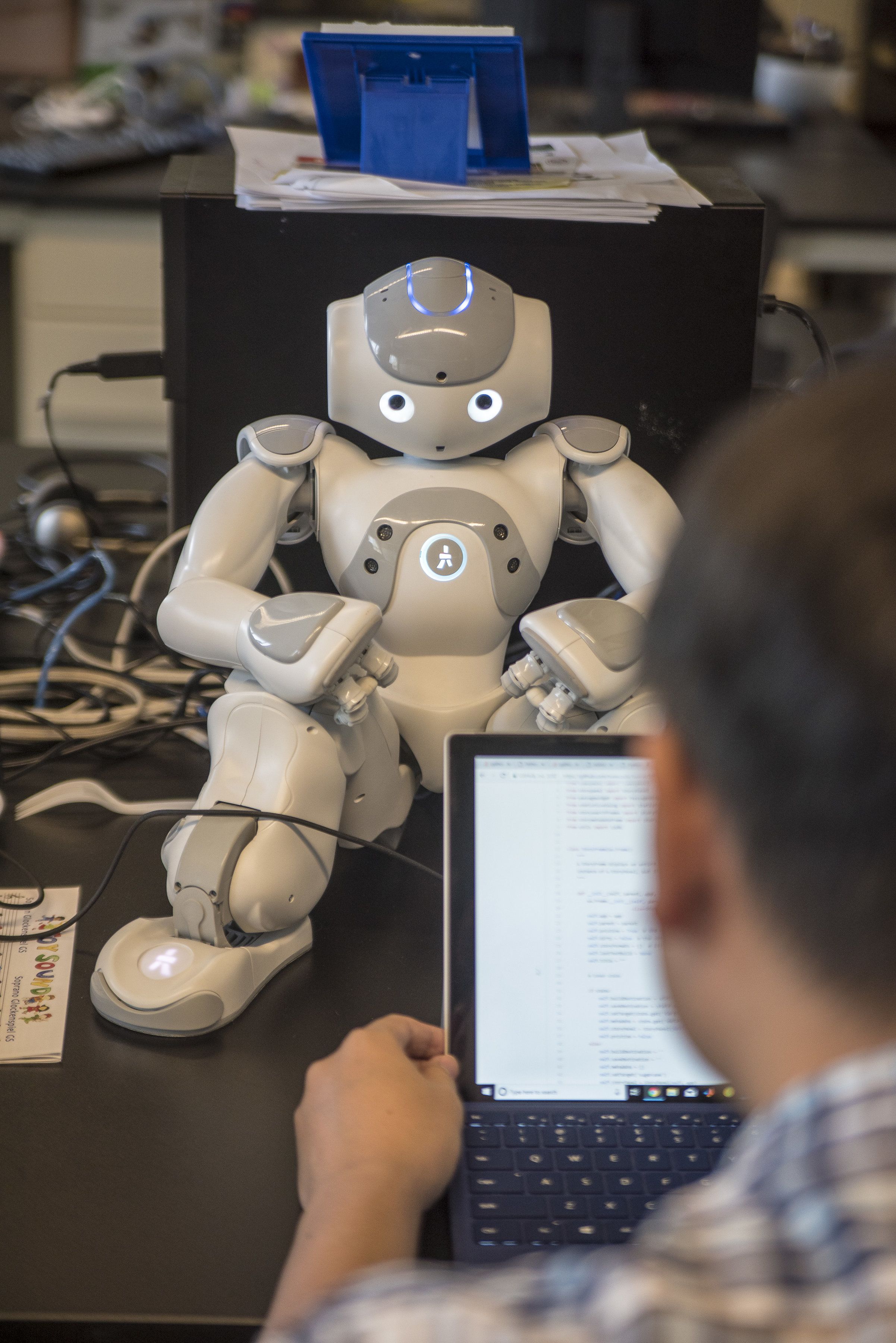 A researcher types code on a laptop screen, with a robot in the background