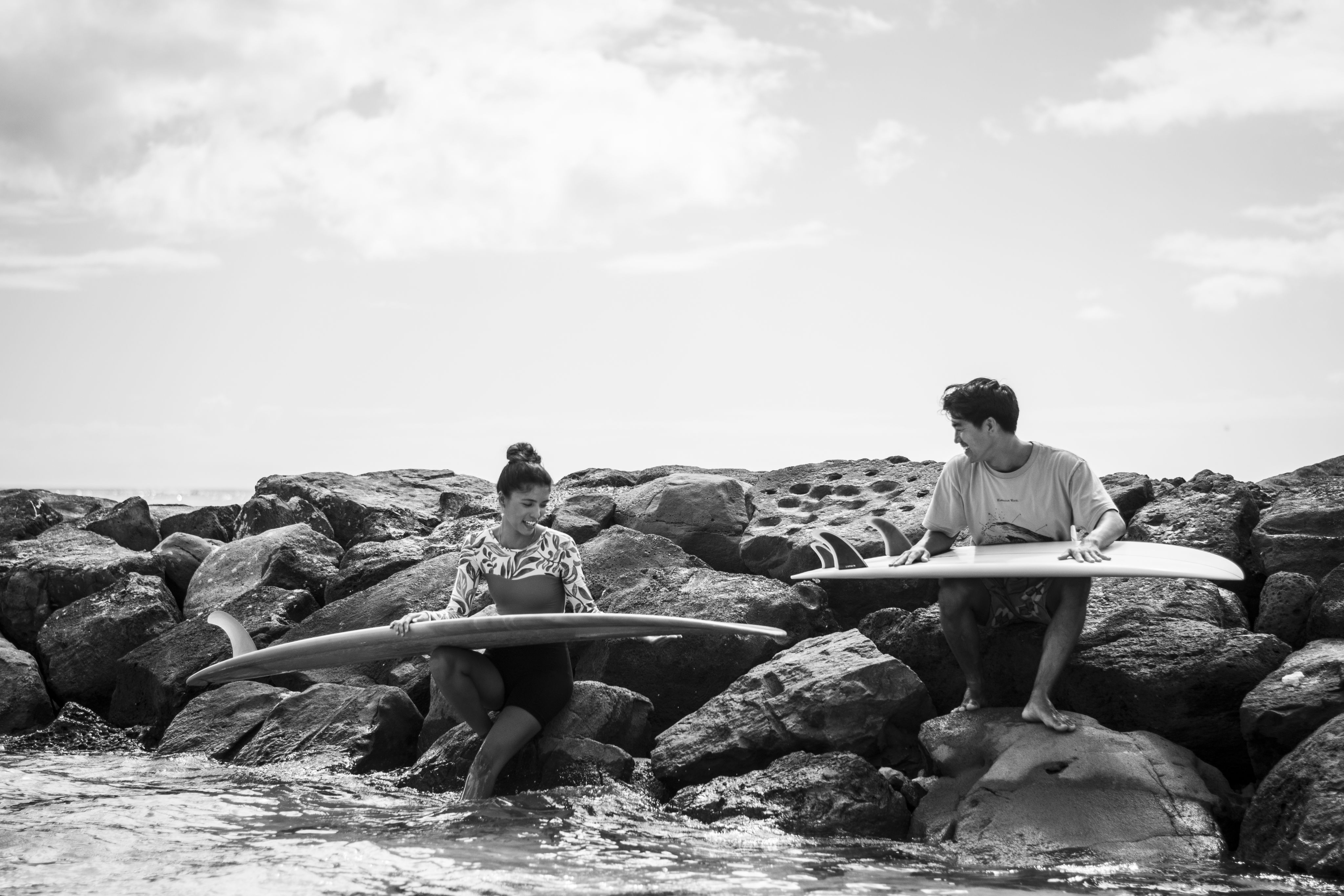 Two surfers sit on the rocks and look down at their boards.