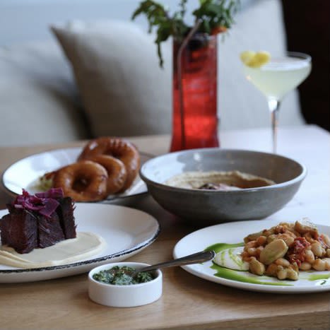 Plates of food and laid out on a table at Zhug. A red glass and a martini glass are behind the plates. A couch with pillows is visible in the background.