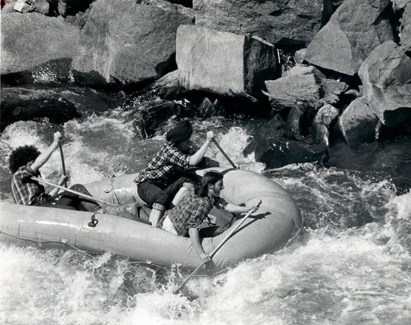 Black-and-white photo of a group of three students white-water rafting along a rushing river.