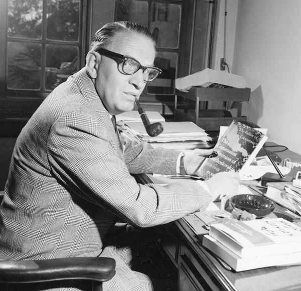 Black and white photo of Joseph Korbel at his desk, holding a book and smoking a pipe.