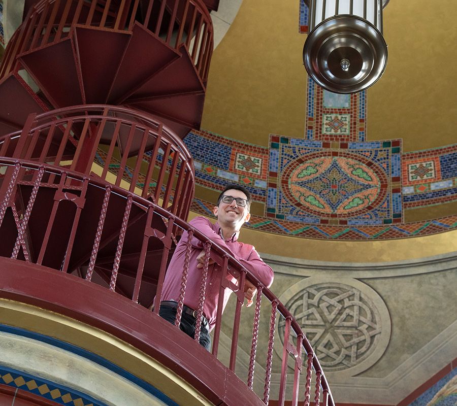 Photo of D-U carillonist Joey Brink standing above us on the red iron spiral staircase that leads to the top of the Carl M. Williams bell tower, intricate tile designs on the wall behind him.