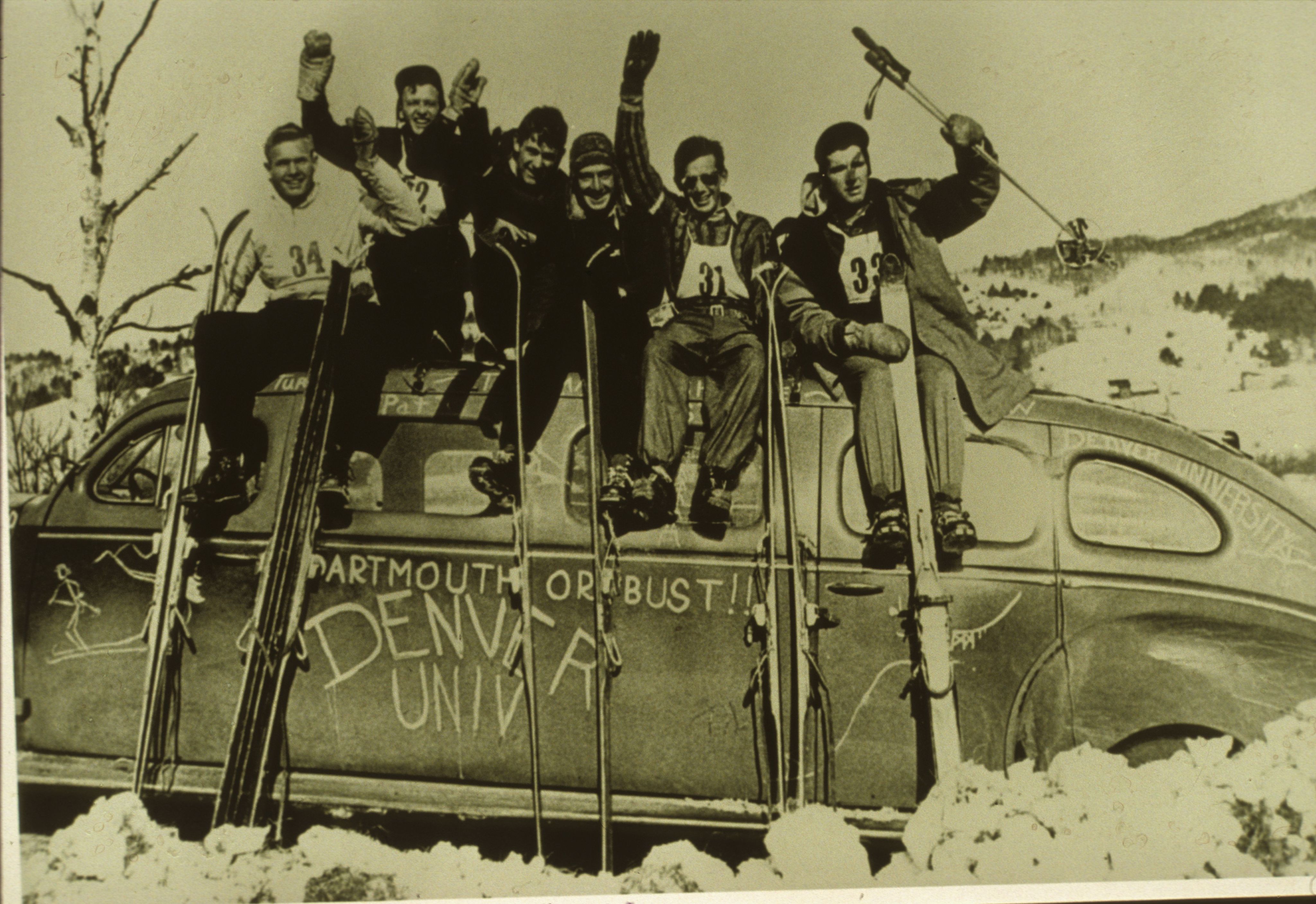 University of Denver ski team members sit on a car that says "Dartmouth or Bust Denver Univ" while holding skis and waving arms in the air. 