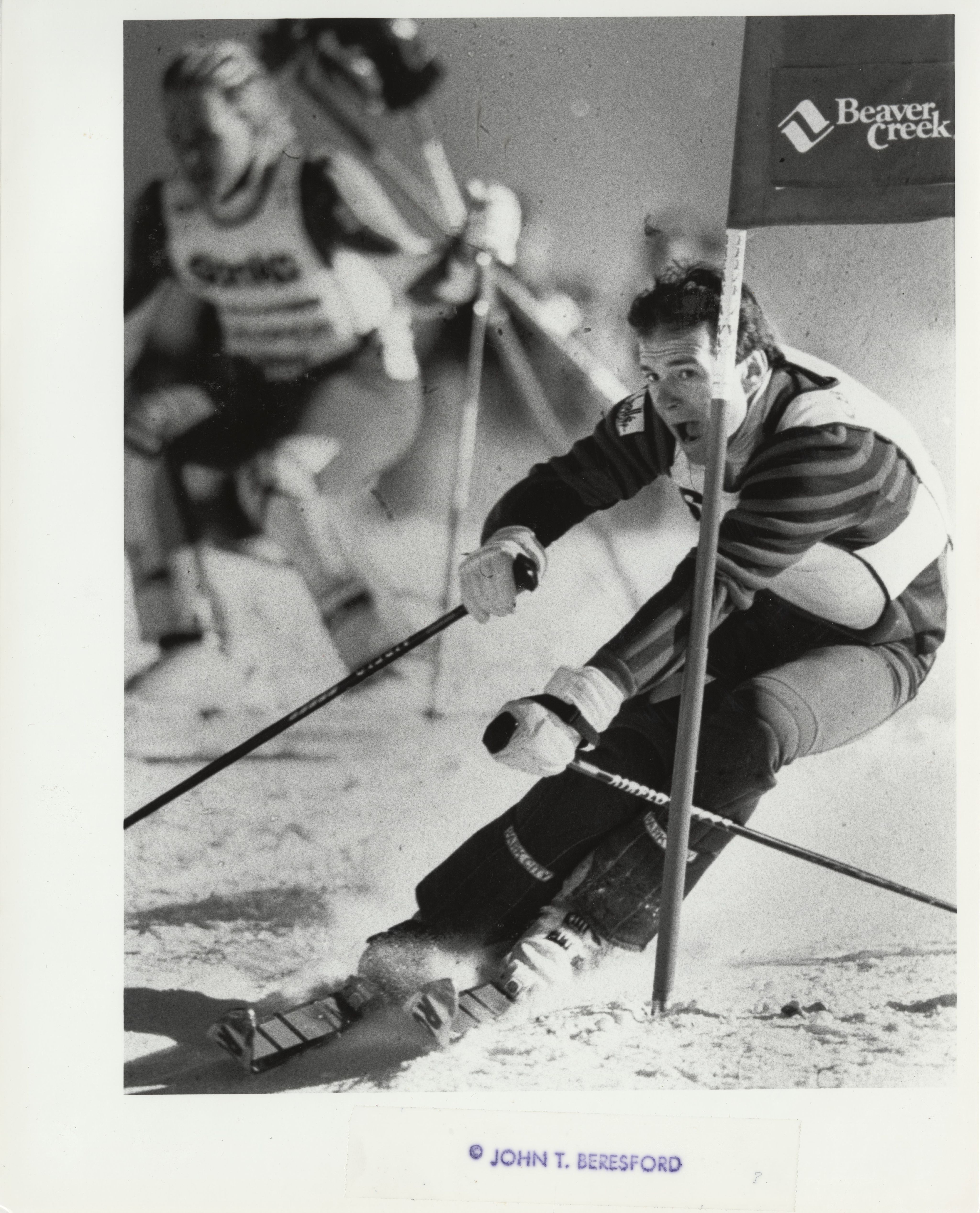 A skier in a race closely rounds the gate flag that reads "Beaver Cree" with his mouth open
