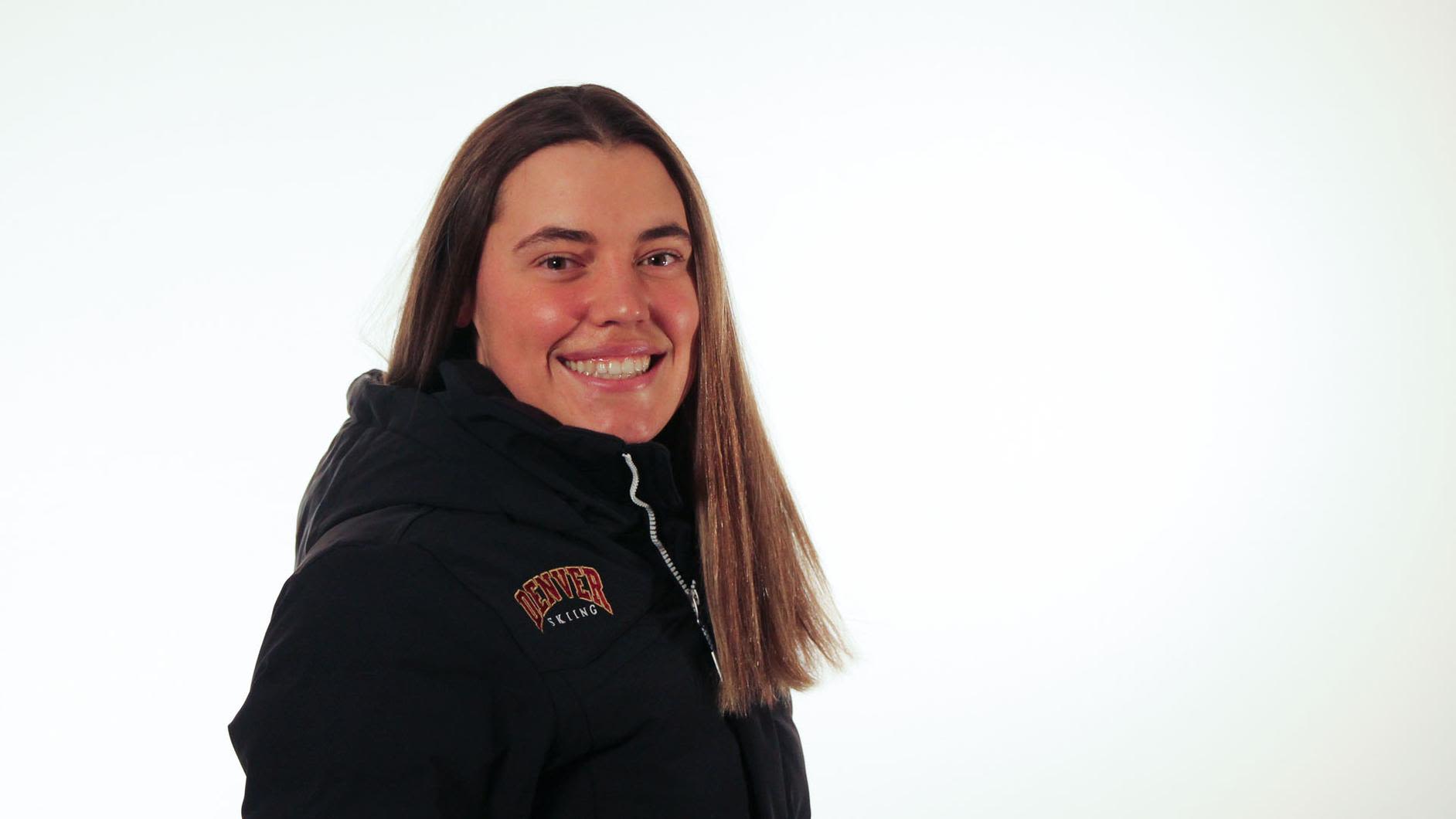A photo of Katie Hensien, wearing a Denver Skiing jacket and smiling at the camera in tront of a plain white background