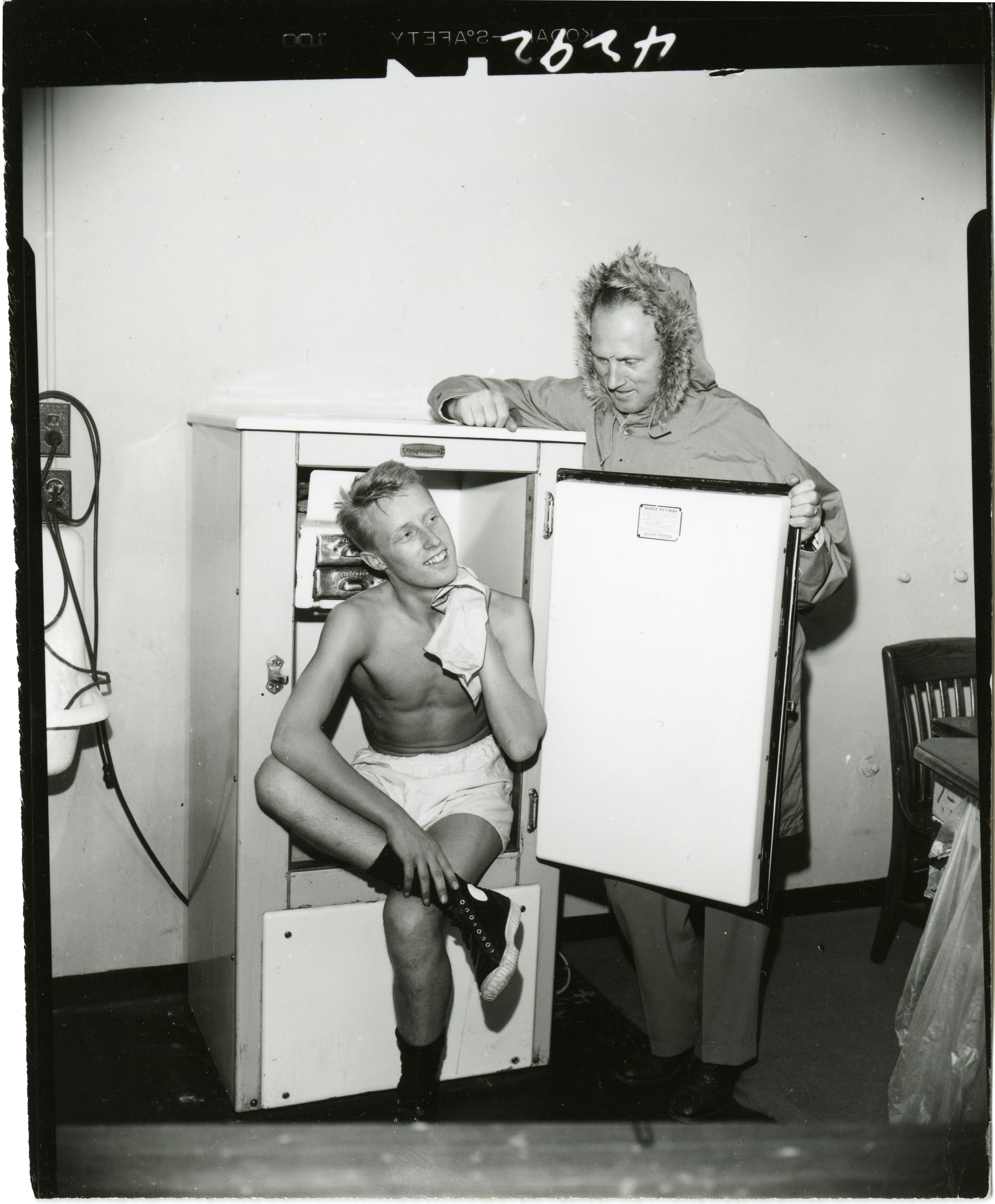 Ski team member in his boxers sits inside refrigerator and wipes his neck with a towel as coach Schaeffler holds the door open and peers down at him 