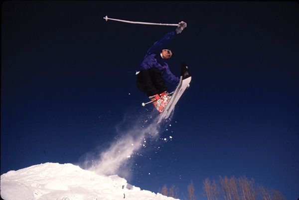 A skier grabs both his skis in one hand and a pole over his head as he jumps off a ski jump