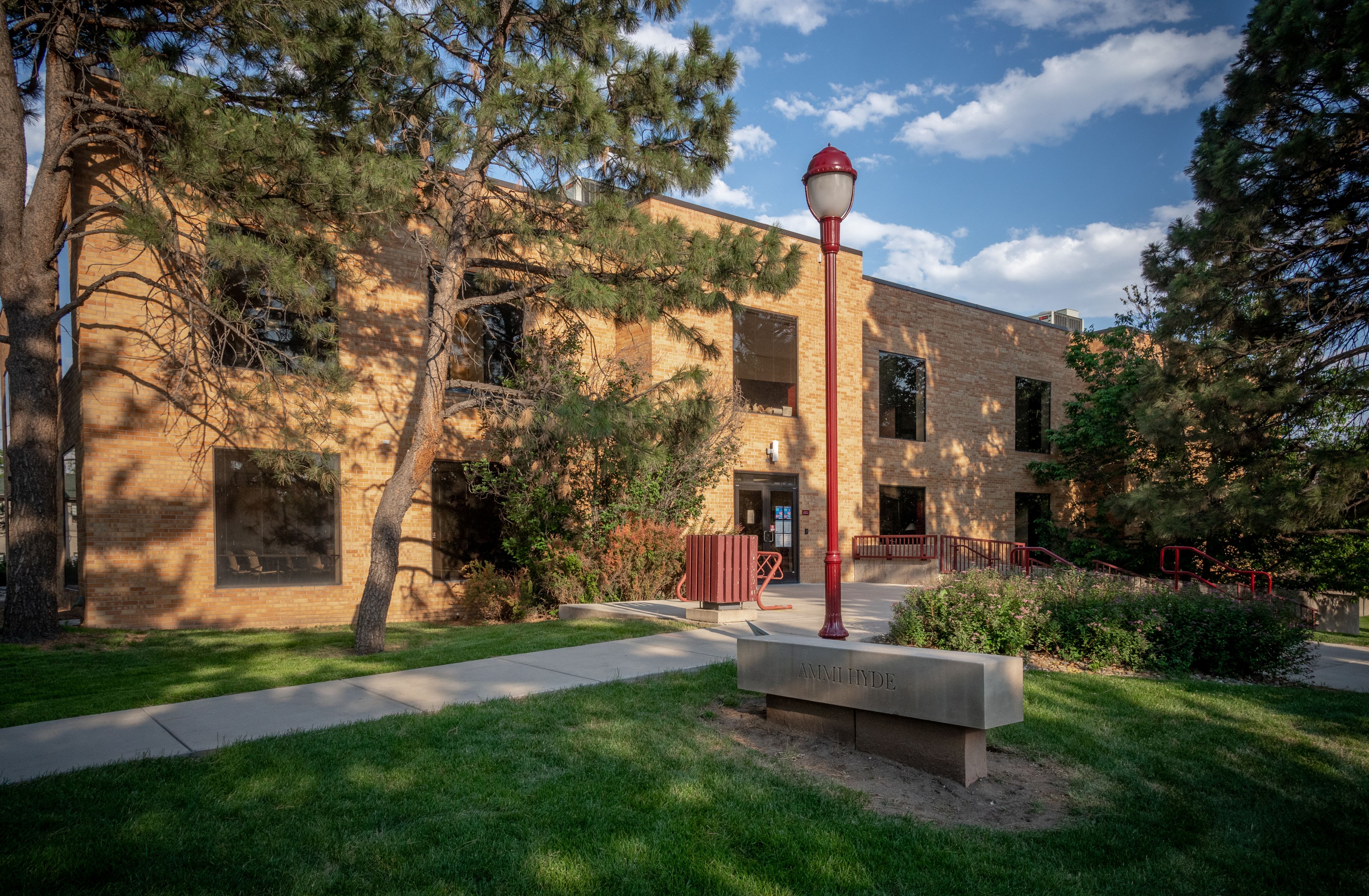 Photo of the front entrance of Ammi Hyde Building, with a lamppost and trees visible in the foreground, a tan building, and blue sky and clouds visible in the background