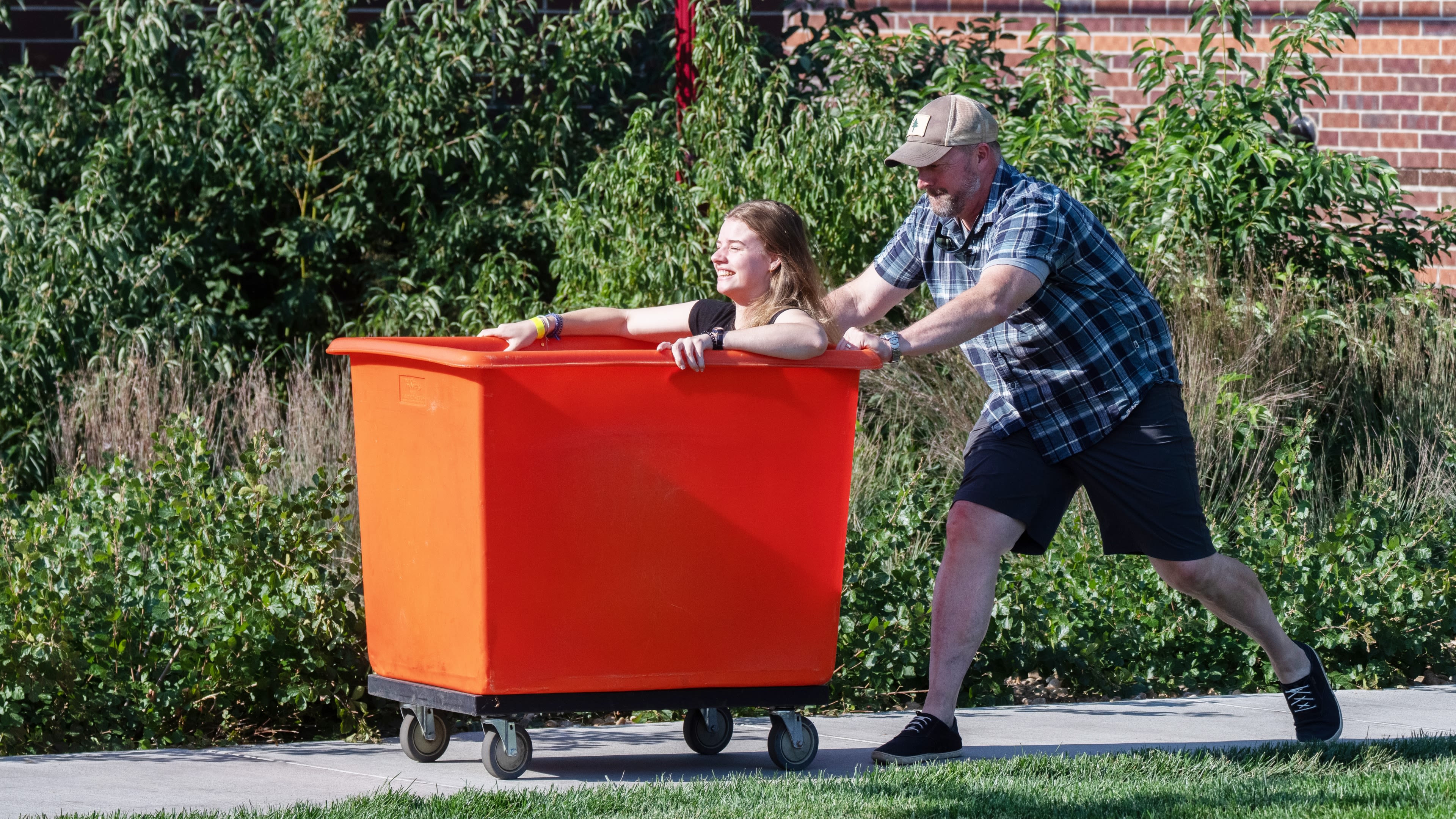 A father pushes his daughter in an orange moving bin across the University of Denver campus 