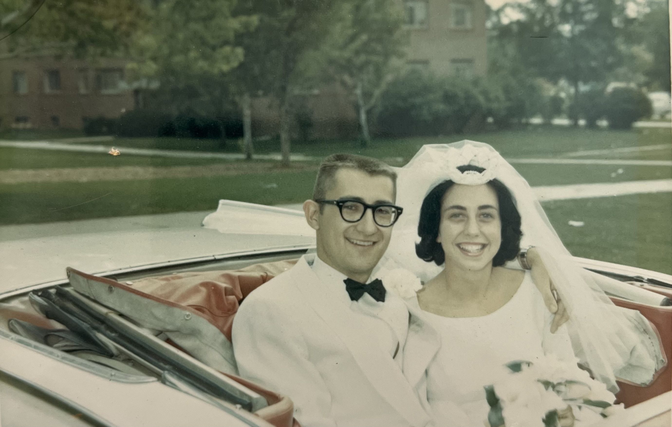 A couple in a wedding dress and suit sit in a car with DU's campus in the background.