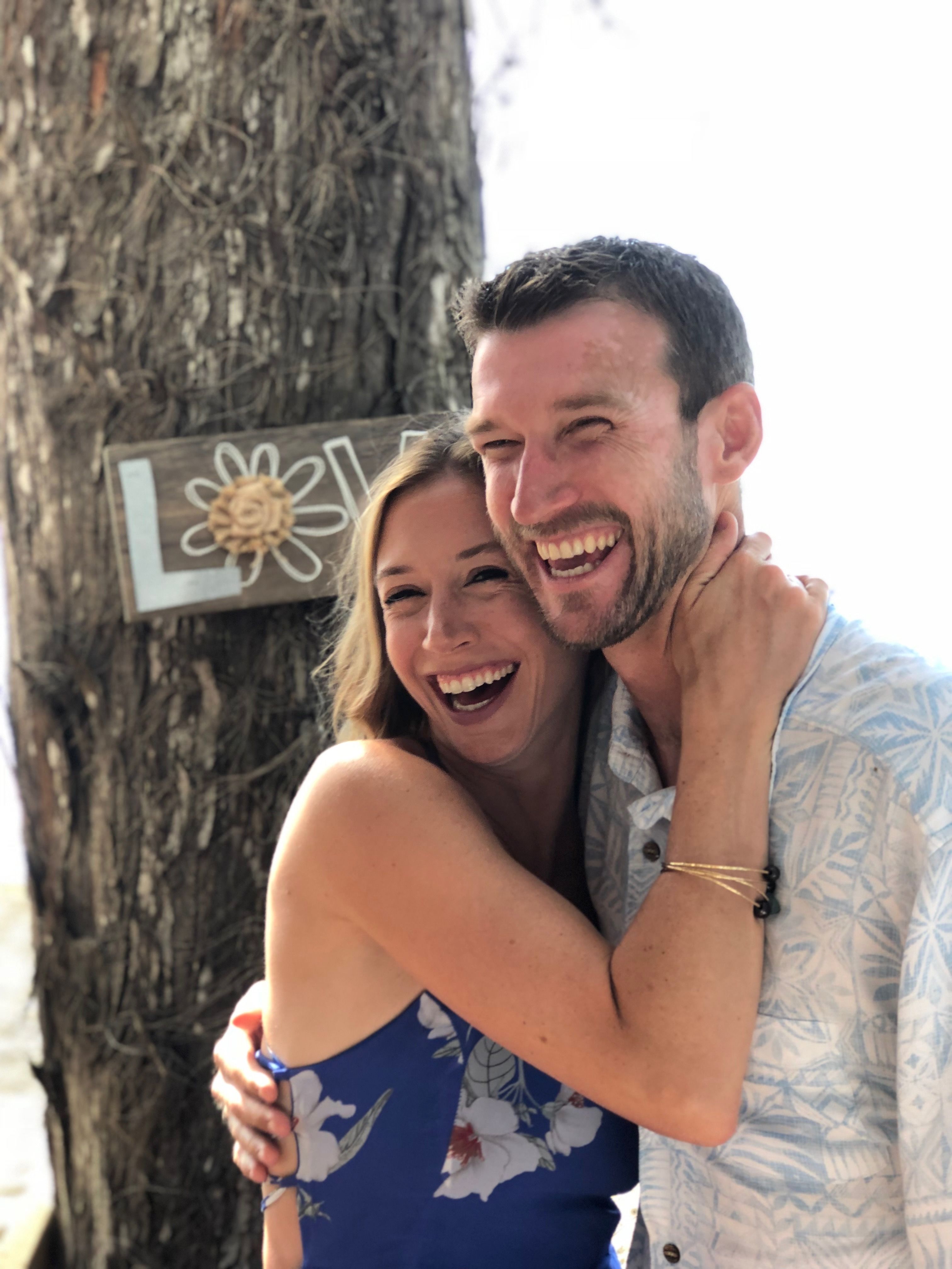 Emily has her arms around Ian in front of a tree as they both smile, looking away from the camera