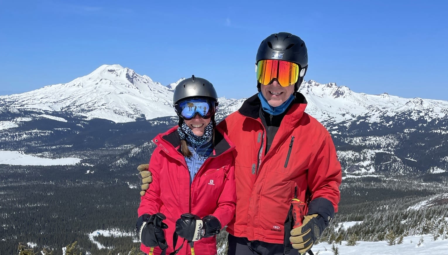 Cathy and Kurt in ski jackets and helmets with the the snow covered rocky mountains in the background