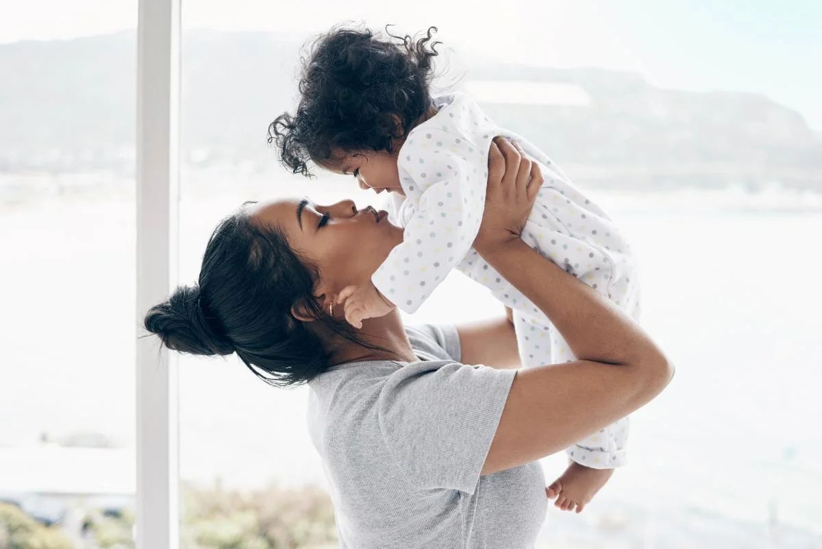a mother in a grey shirt holds up a baby in a white onesie