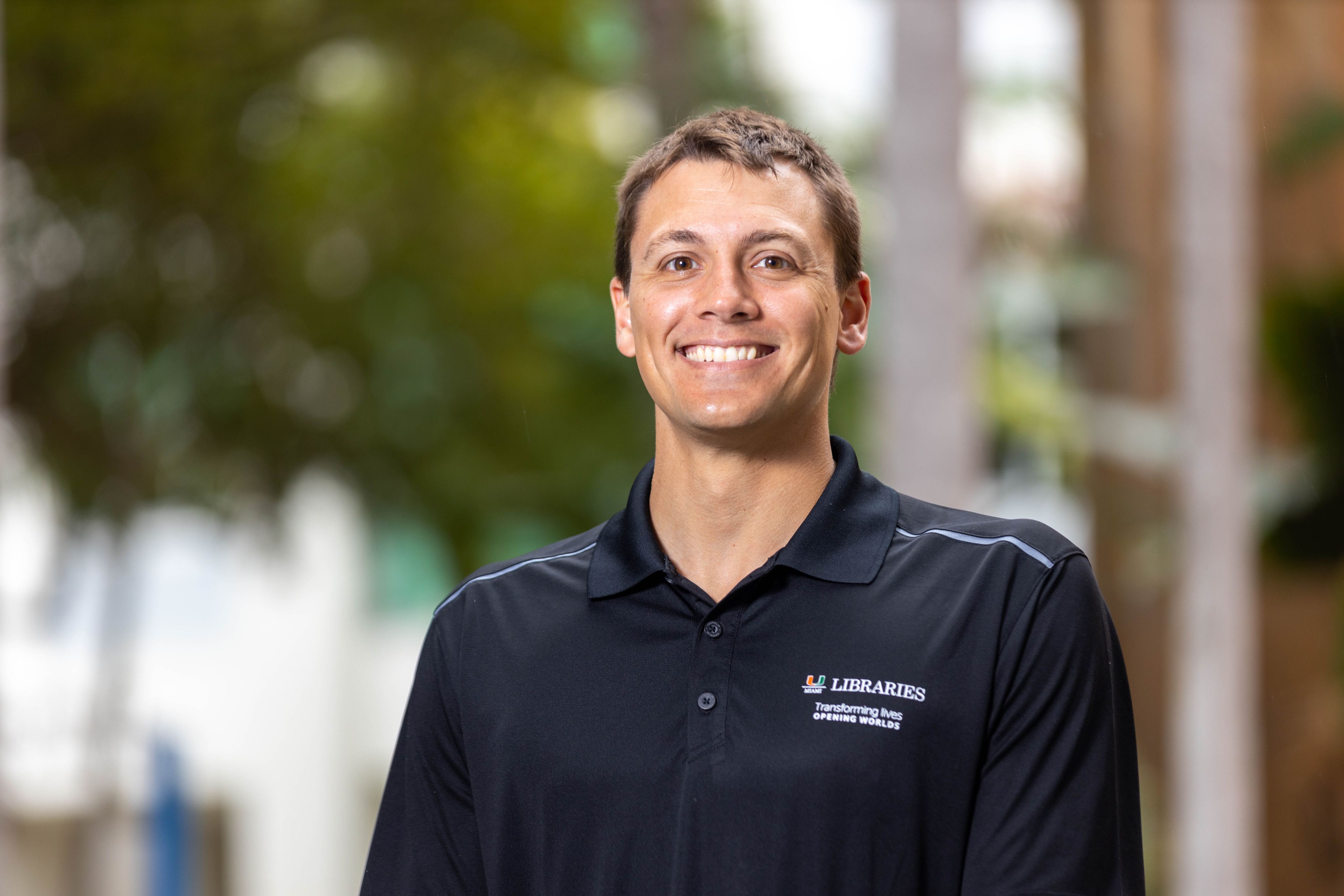 Nick Iwanicki in a Miami Libraries shirt smiles for a professional photo with the greenery behind him blurred