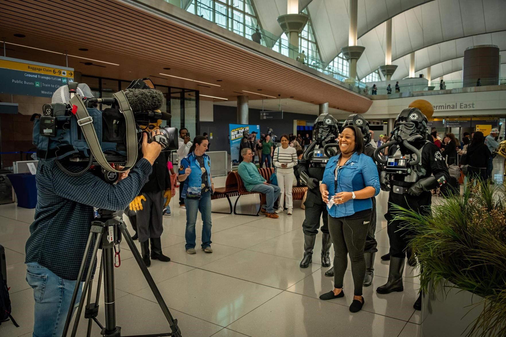 Ashely stands in Denver International Airport in front of a broadcast camera with a crowd behind her. 