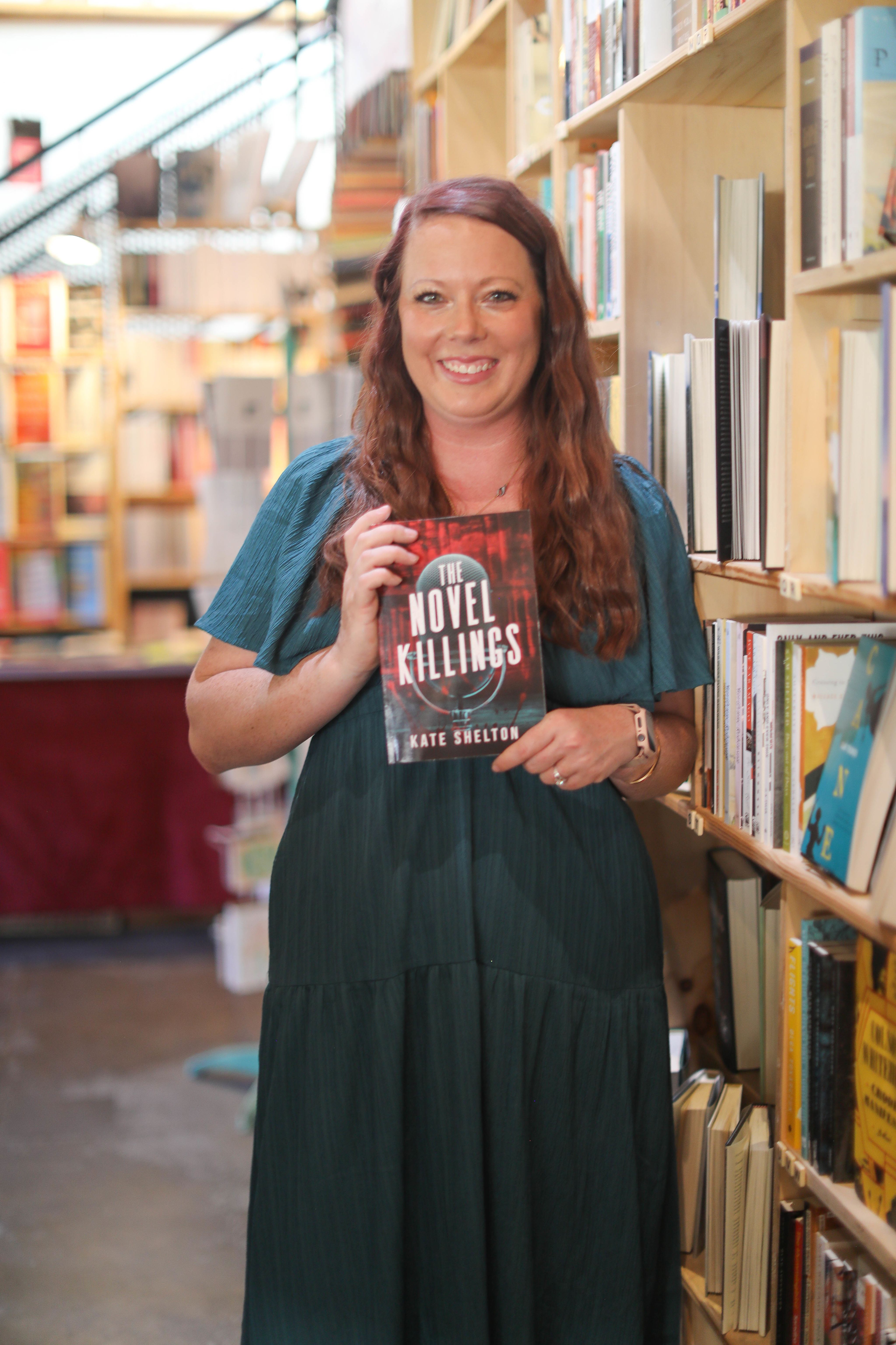 Kate Shelton in a library, holding a copy of her debut book.