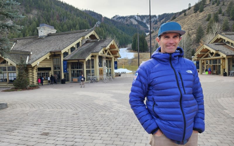 Stephen Helfenbein poses in a blue coat in front of rustic-looking buildings amid a mountain range.