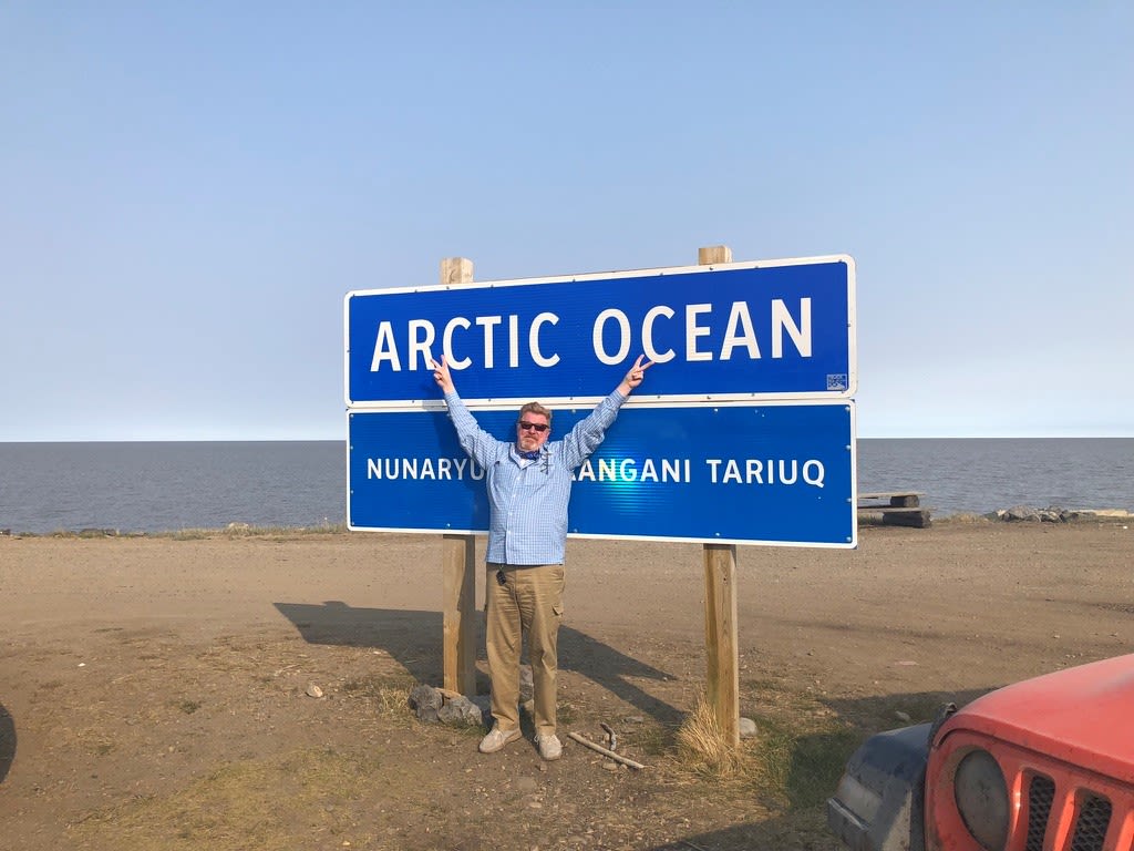 Photo of Tom Cryer standing with his arms outstretched over his head in front of a large blue sign that says "Atlantic Ocean" in all caps.