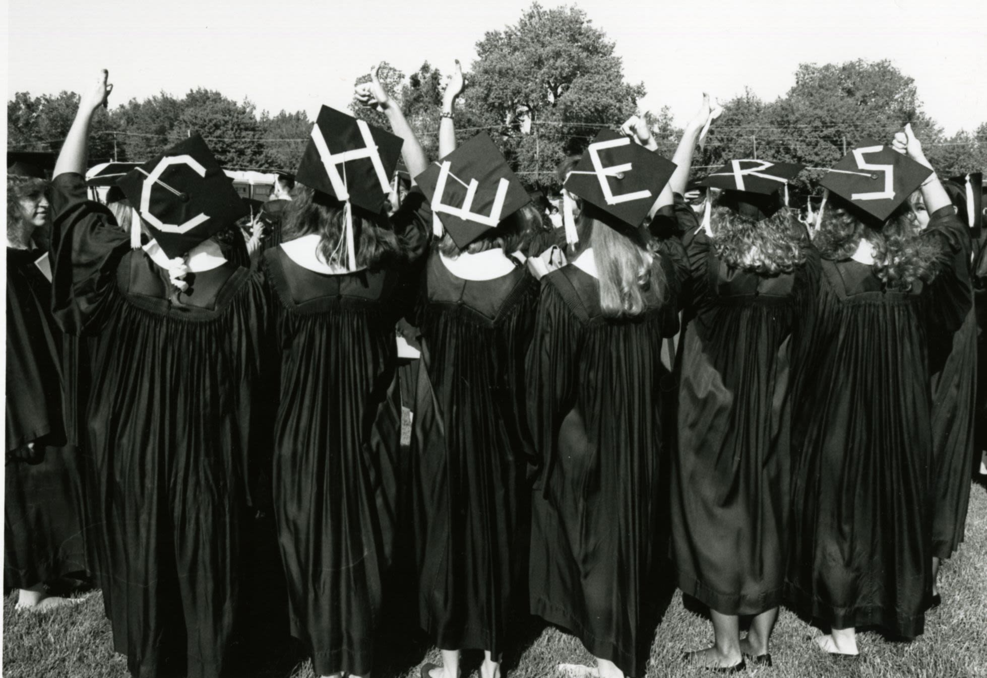 A group of graduates face away from the camera with letters written on their graduation caps that when in a line, spell "Cheers" 